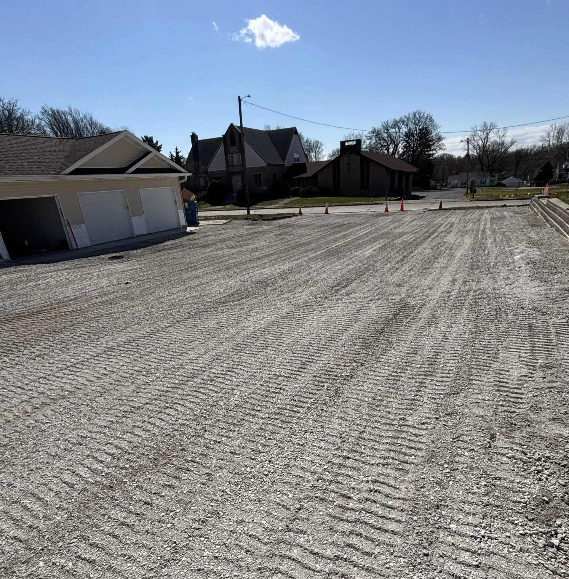 Gravel road under construction, houses in the background, blue sky.