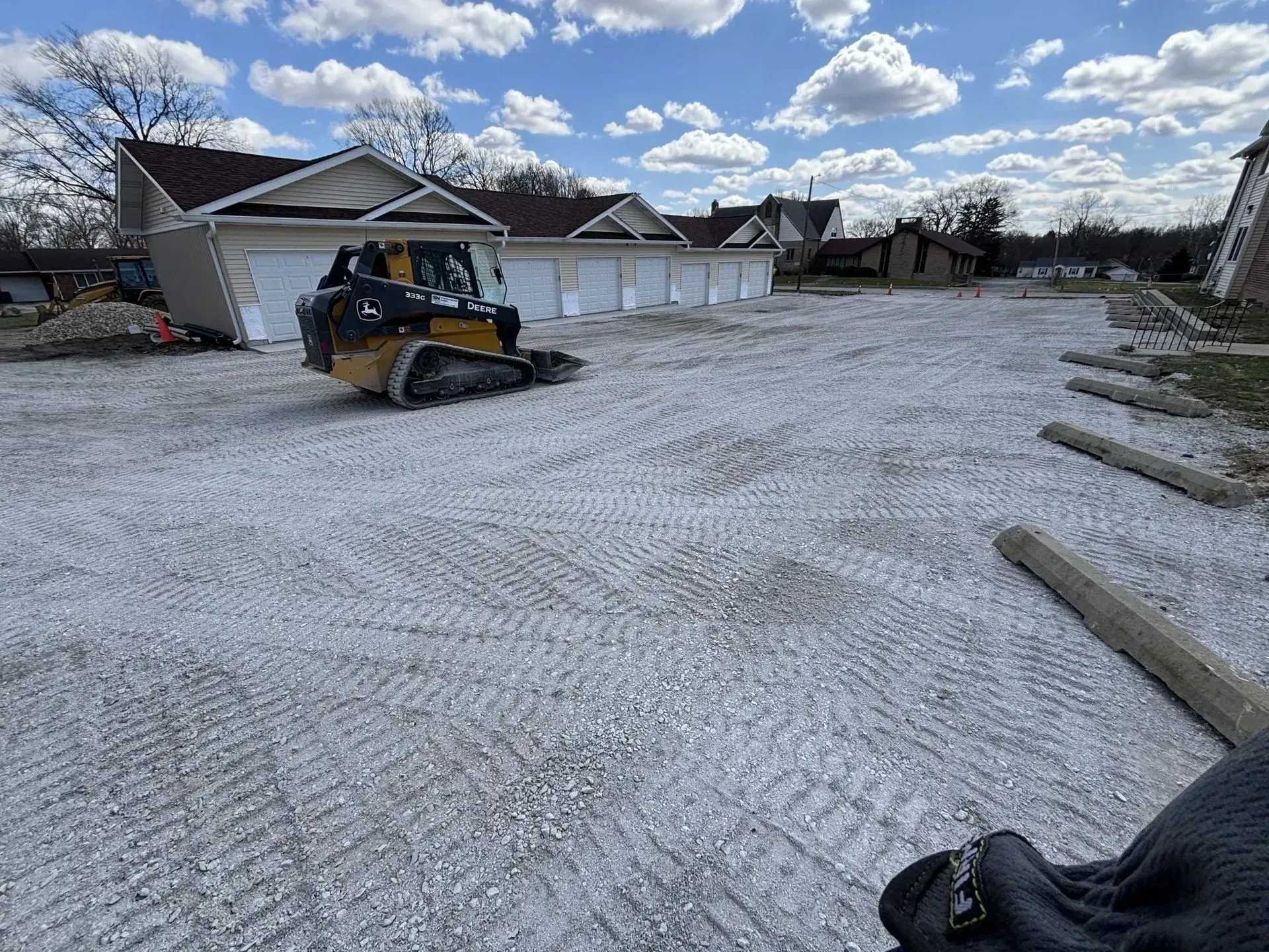 A small construction vehicle smoothing gravel in front of white garages on a sunny day.