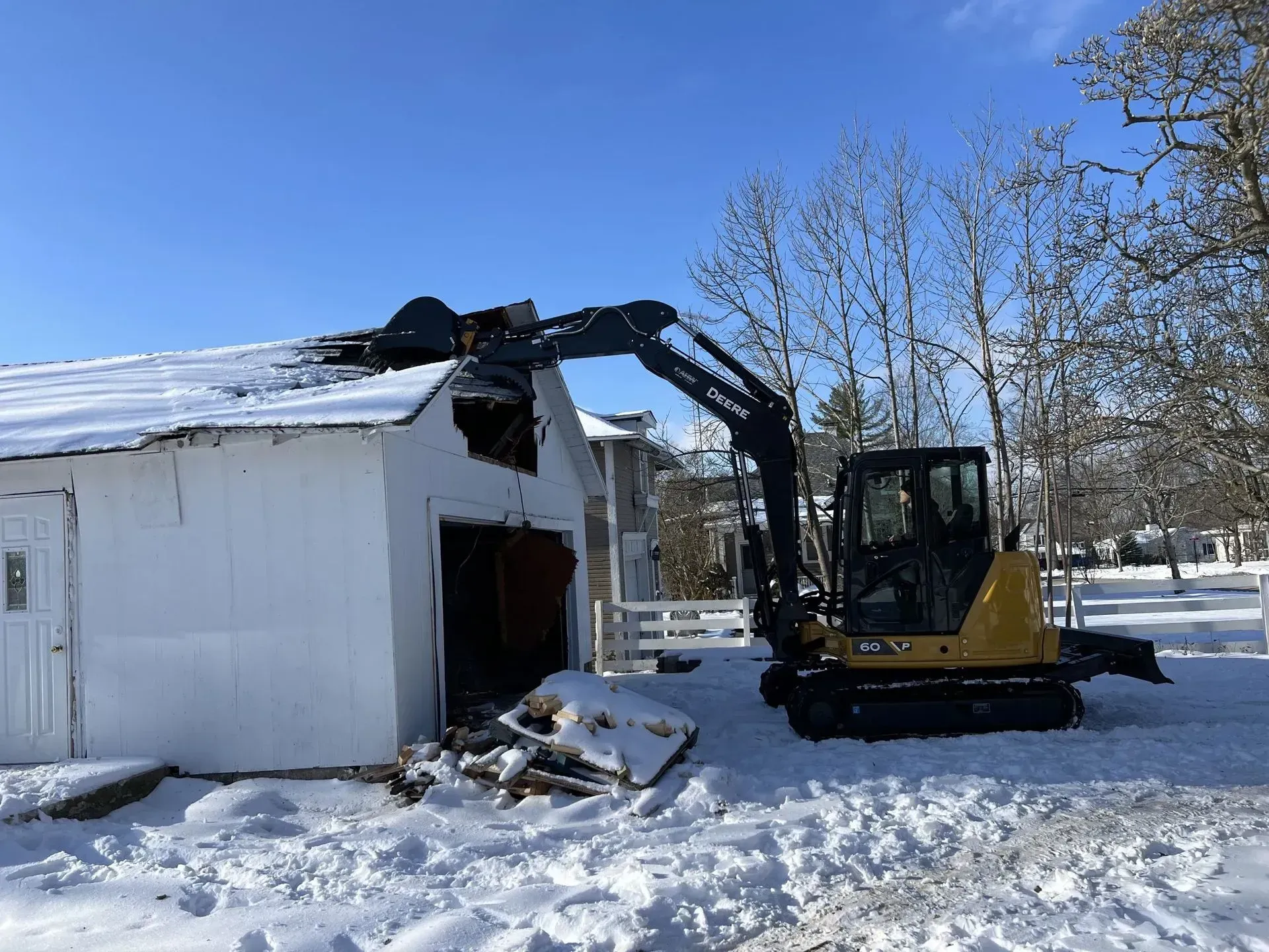 A small excavator demolishing a white building with a snow-covered roof.