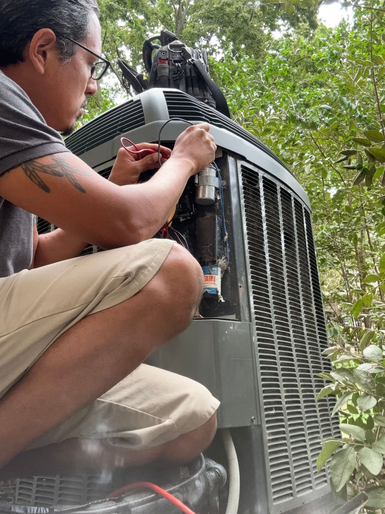 Man in glasses working on an air conditioner unit outdoors, surrounded by greenery.