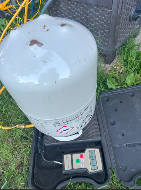 White propane tank on a scale with a digital display outdoors on grass.