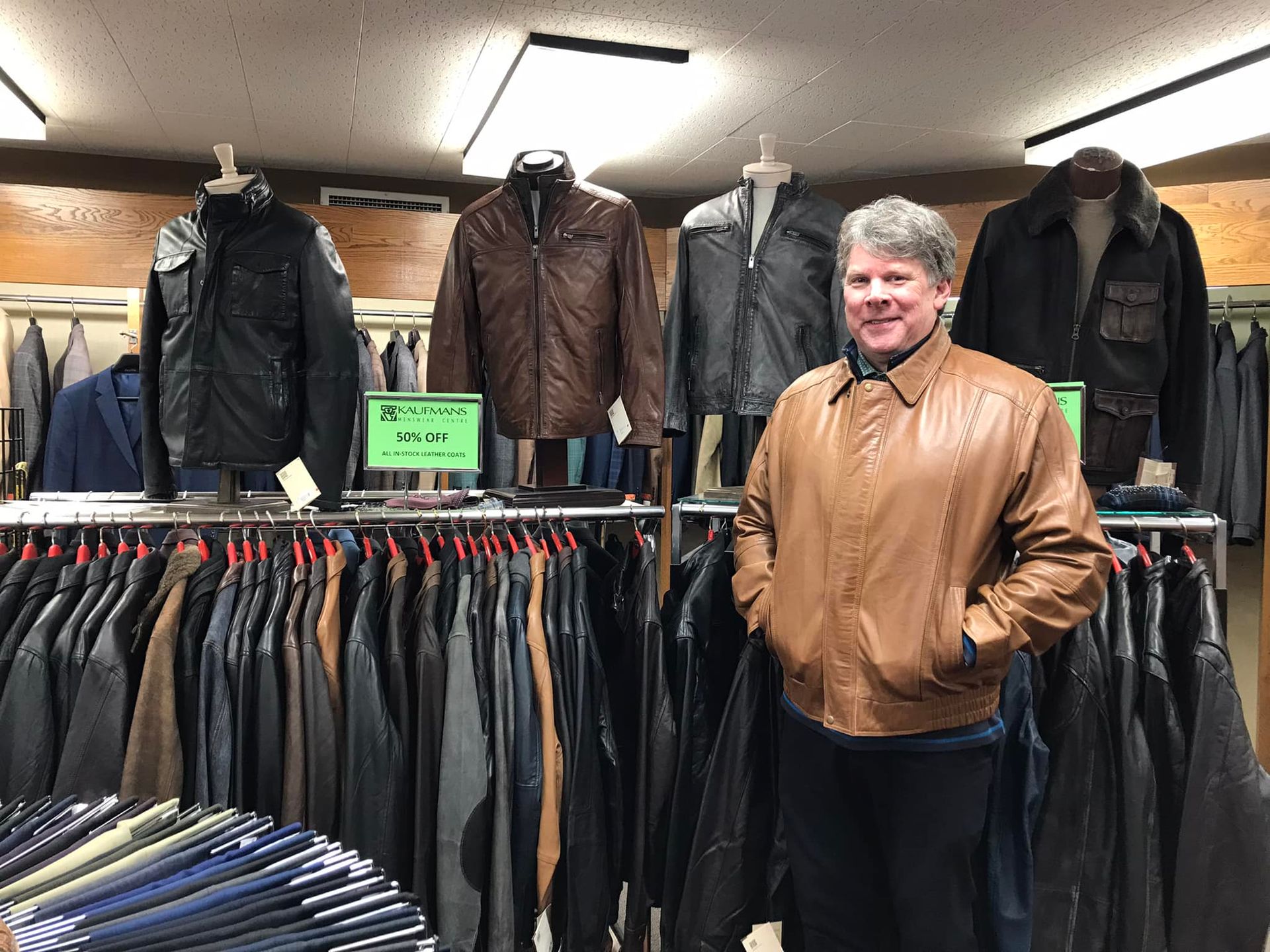 Man in brown leather jacket stands in a clothing store, surrounded by jackets on display.