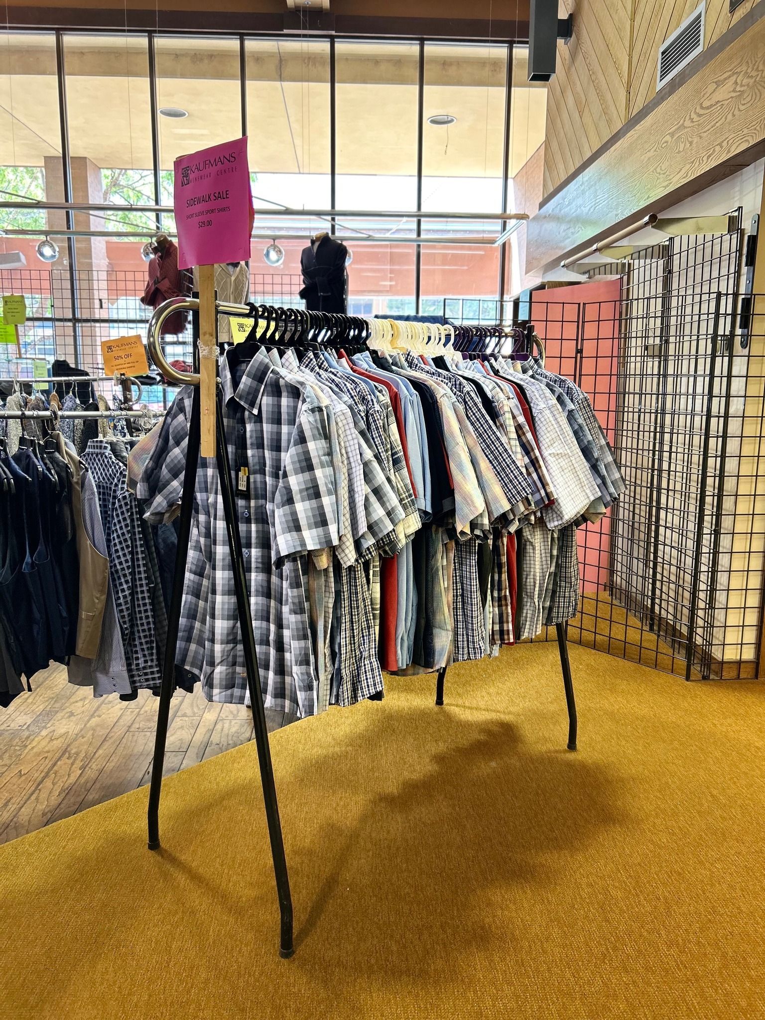 Clothing rack with shirts on display, inside a building. Pink sign atop a wooden post. Yellow carpet.
