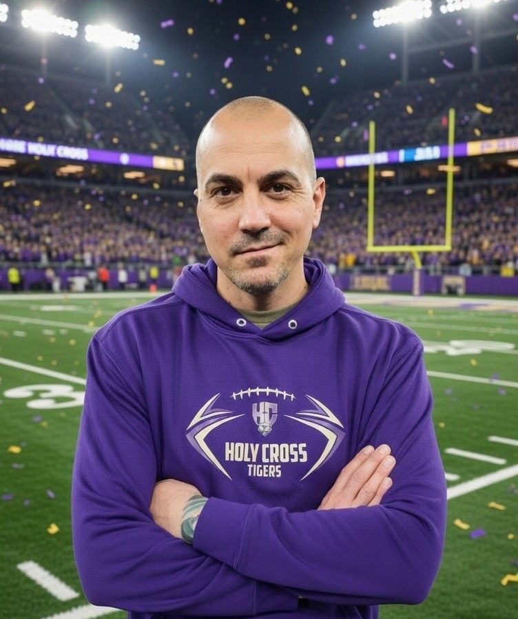 Man with crossed arms on a football field. He is wearing a blue polo shirt, khaki pants, and has a serious expression.