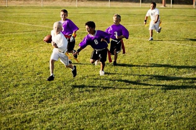 Flag football game at night, a player in red jersey runs with the ball as others pursue on a green field.