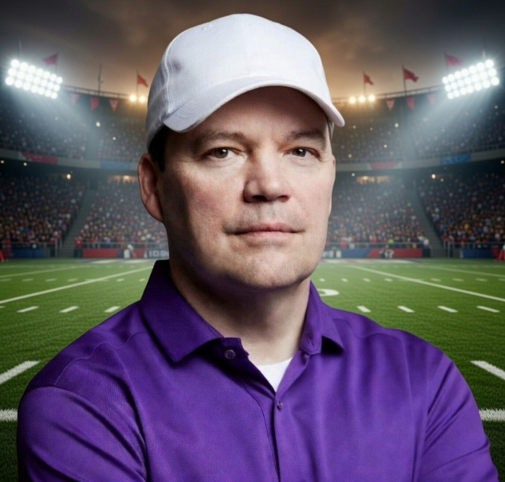 Football coach, arms crossed, on the field. Blue shirt, serious expression, players in background.