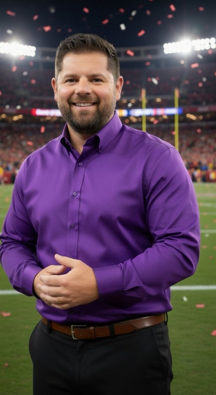 Man with crossed arms on a football field. He is wearing a blue polo shirt, khaki pants, and has a serious expression.