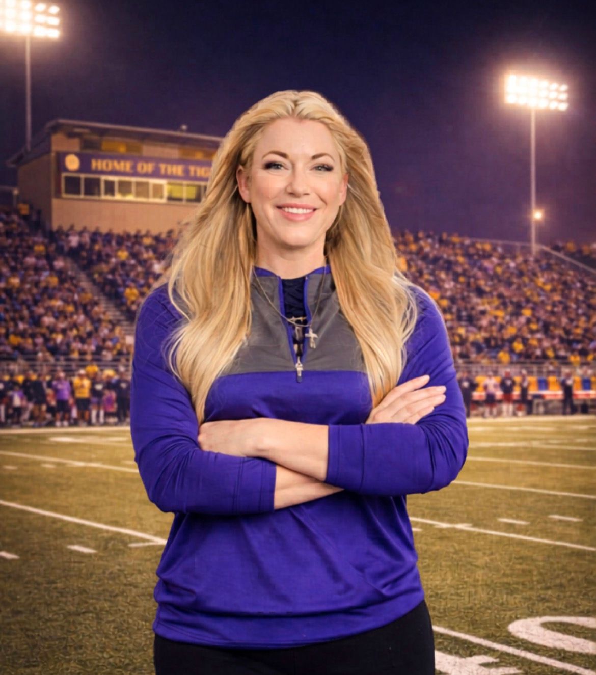 Football coach, arms crossed, on the field. Blue shirt, serious expression, players in background.