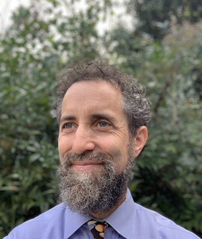 Man with a beard and blue shirt, looking towards the right, outside with greenery in the background.