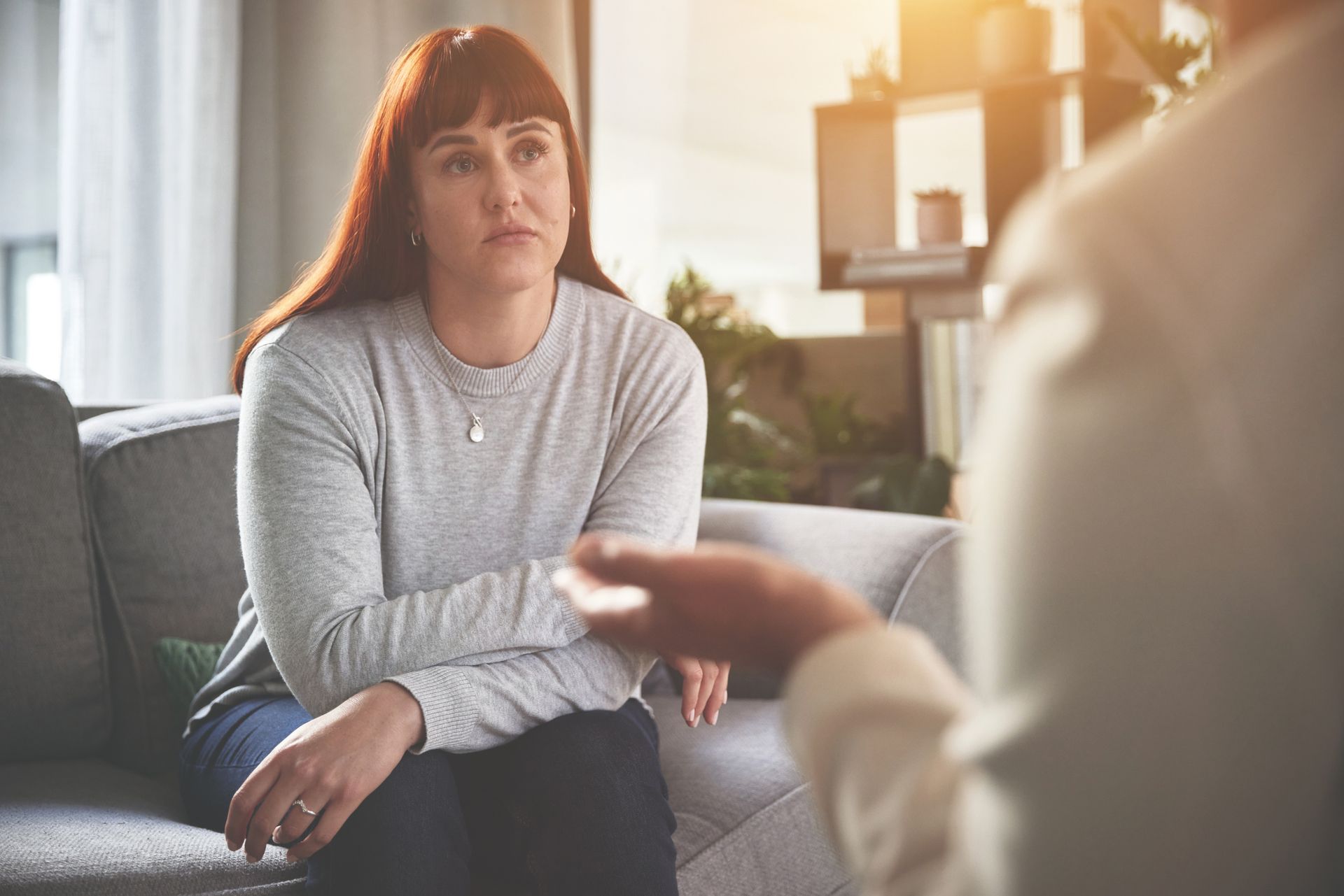 Woman with red hair, gray shirt, in therapy session on a sofa, listening to a person.