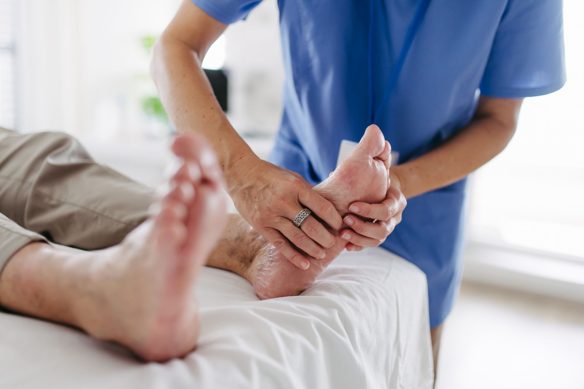 Nurse in blue scrubs massaging the foot of a person lying on a white bed.