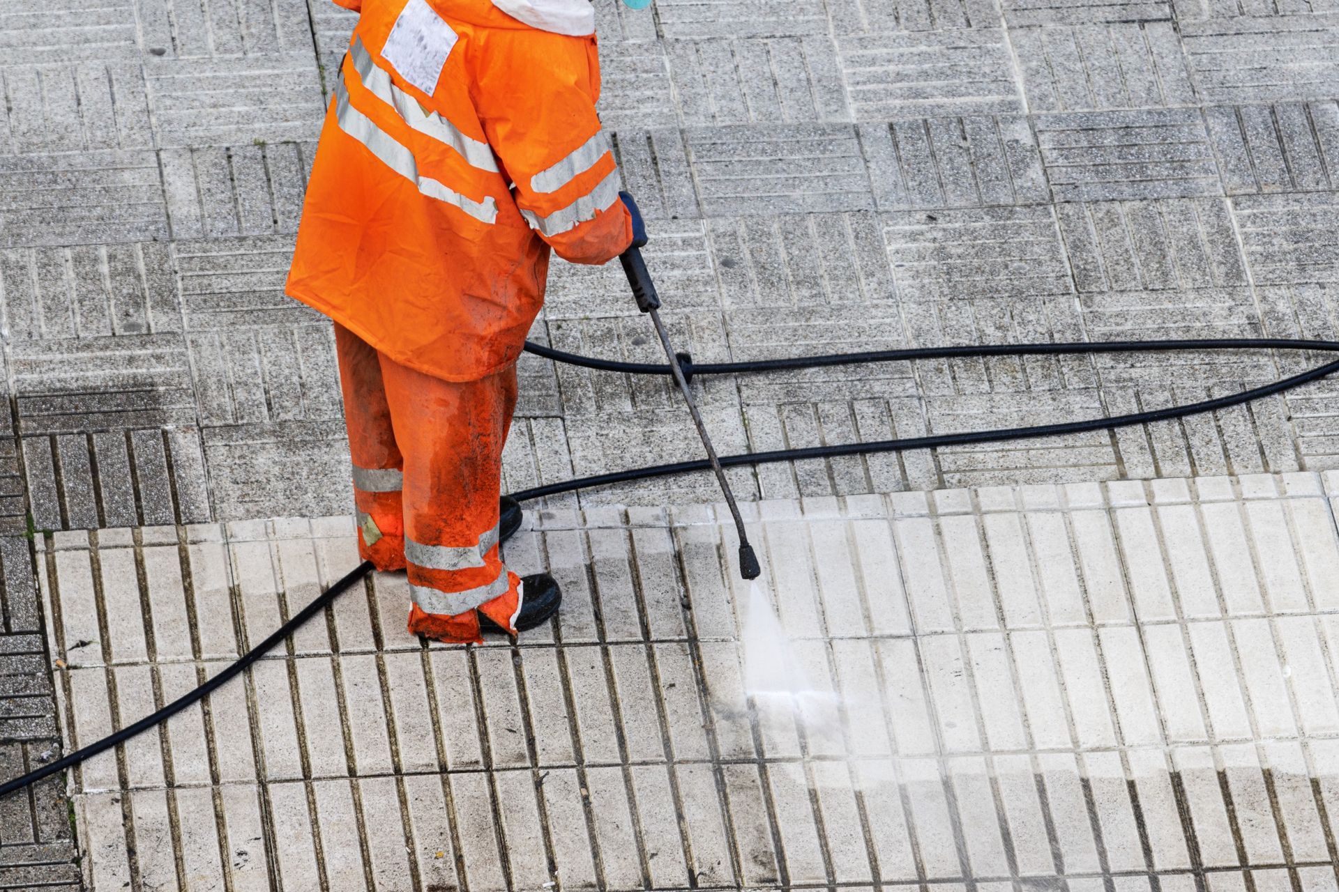 A man in an orange jacket is cleaning a tiled floor with a high pressure washer.