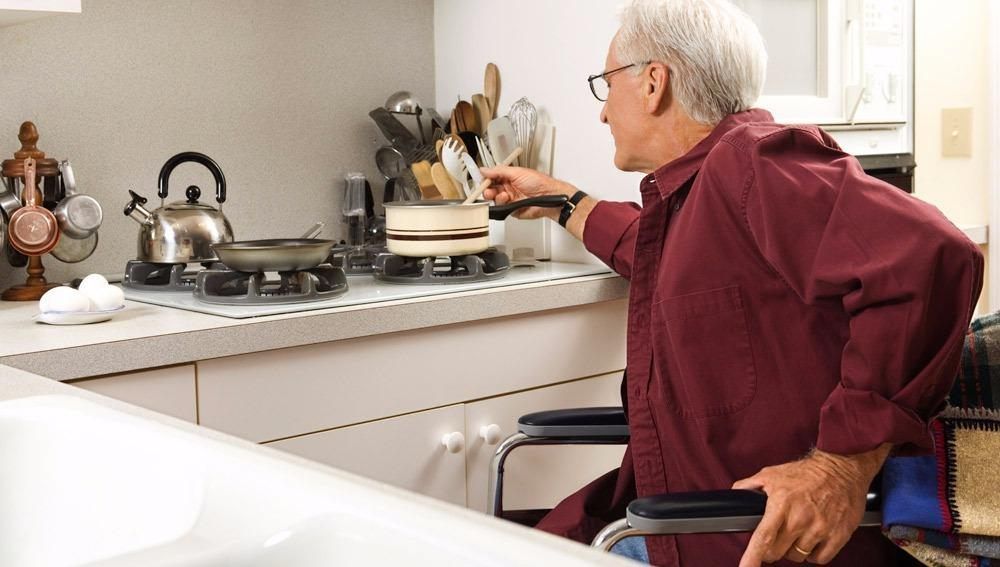 An older man in a wheelchair is cooking in a kitchen.