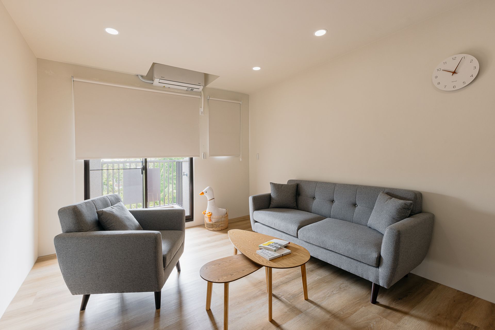 客廳裡有灰色的沙發和扶手椅、兩張小木桌、有百葉窗的窗戶和一塊鐘。