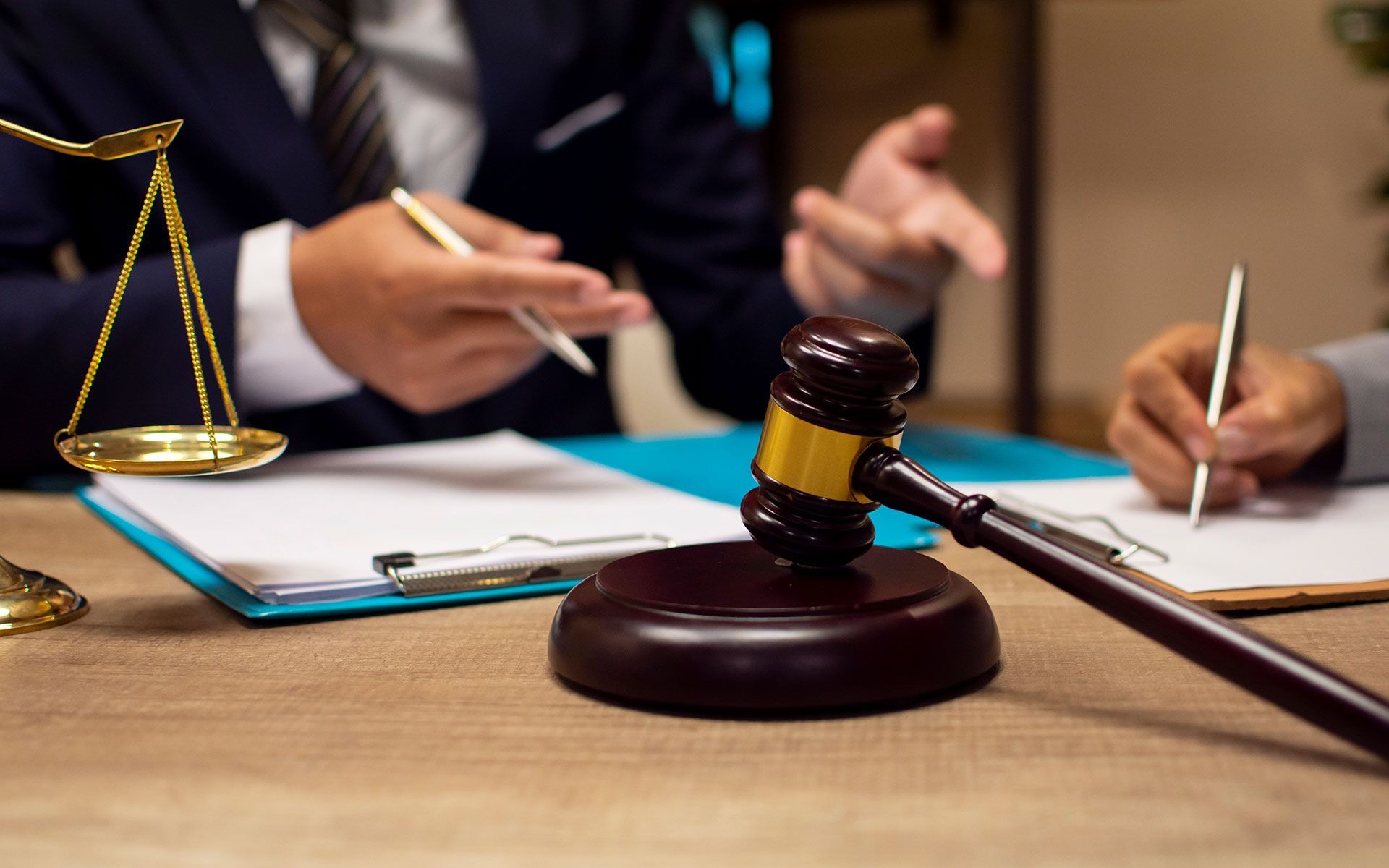 Gavel on a wooden desk with a scale and legal documents, two people in suits, one gesturing, the other writing.