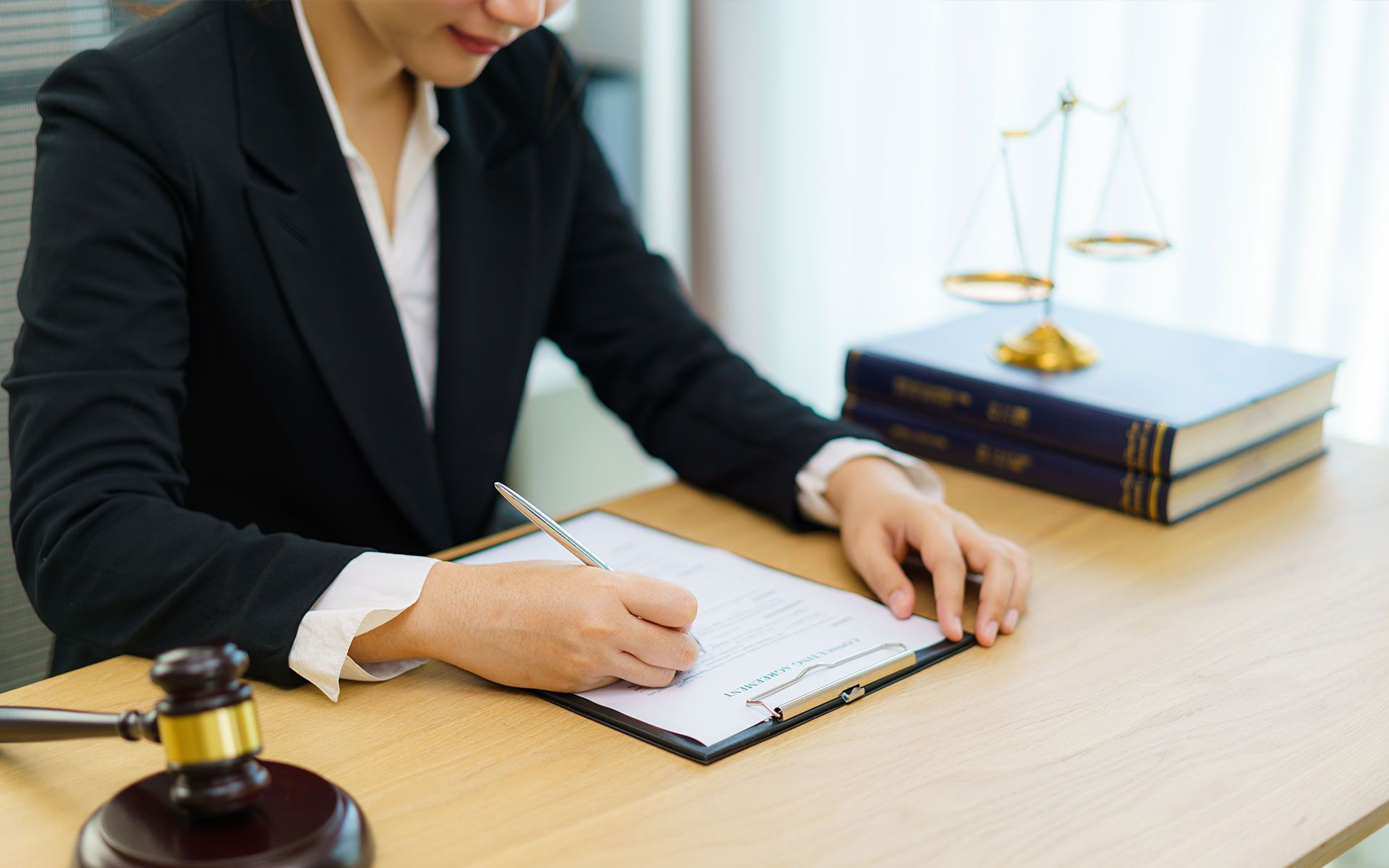 Lawyer in a black suit writing on a clipboard at a desk; scales of justice and law books in background.