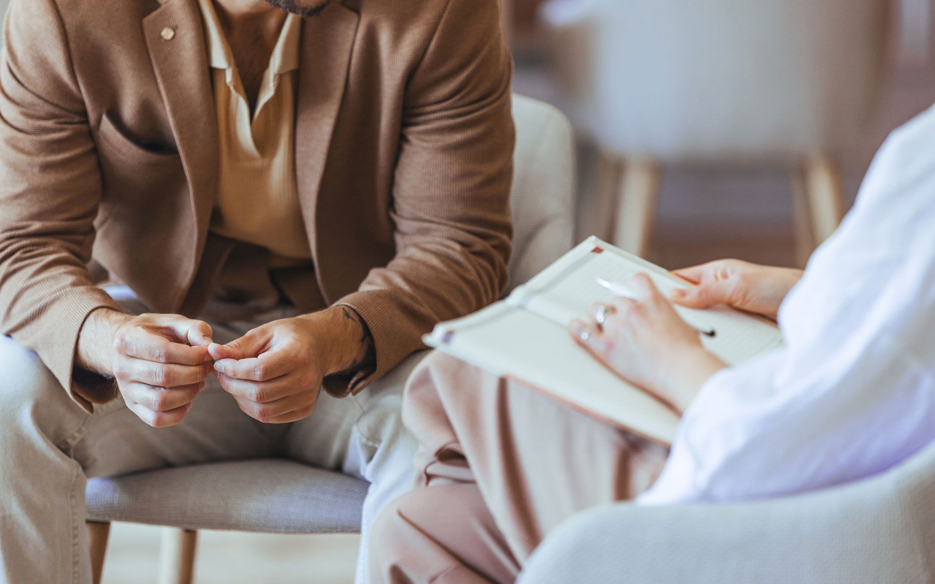 Person in a brown jacket and tan pants seated, consulting with another person holding a notepad.