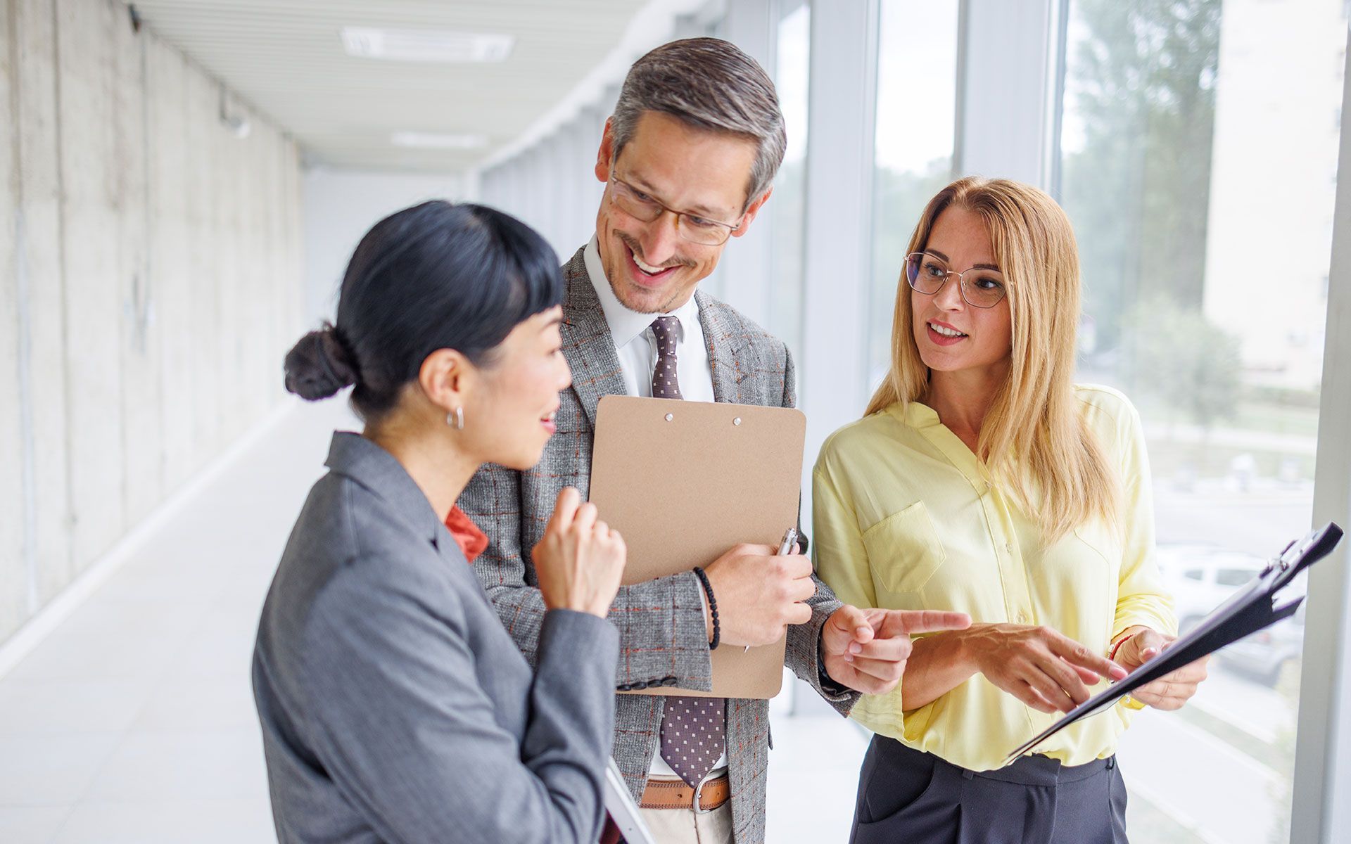 Three businesspeople in a hallway, discussing documents. Man holding a clipboard smiles at two women.