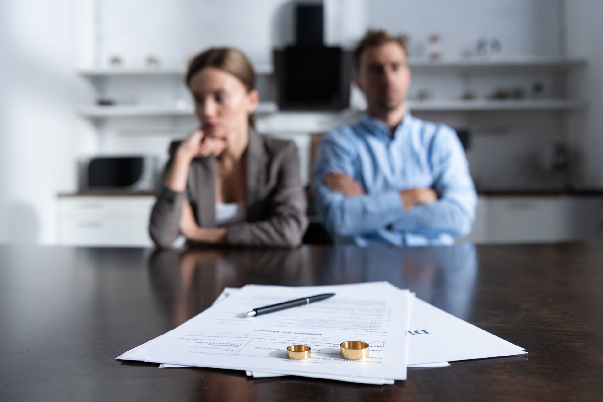 Divorce papers and wedding rings on a table with two people sitting apart in the background.