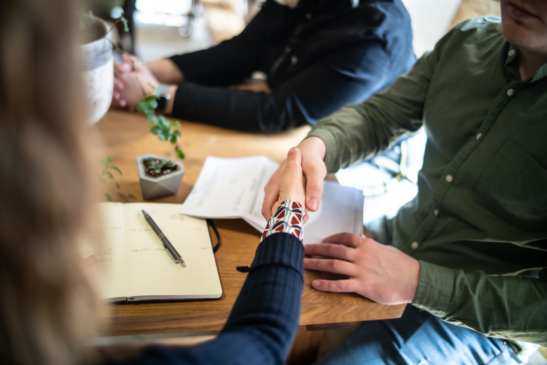 Two people shake hands over a wooden desk with a notebook and a small plant, while a third person sits nearby.