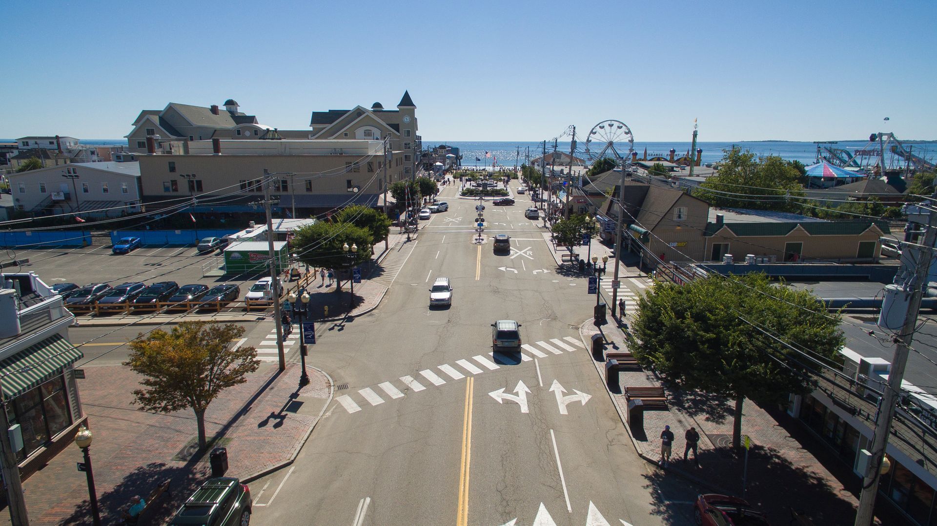 An elevated street view of a sunny coastal town with shops, cars, and a distant amusement park pier.