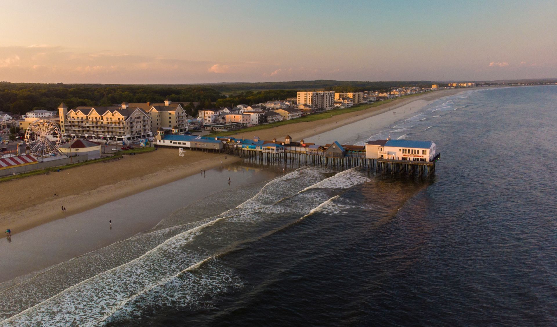 Aerial view of Old Orchard Beach pier extending into the Atlantic Ocean at sunset, with coastal buildings and sand.