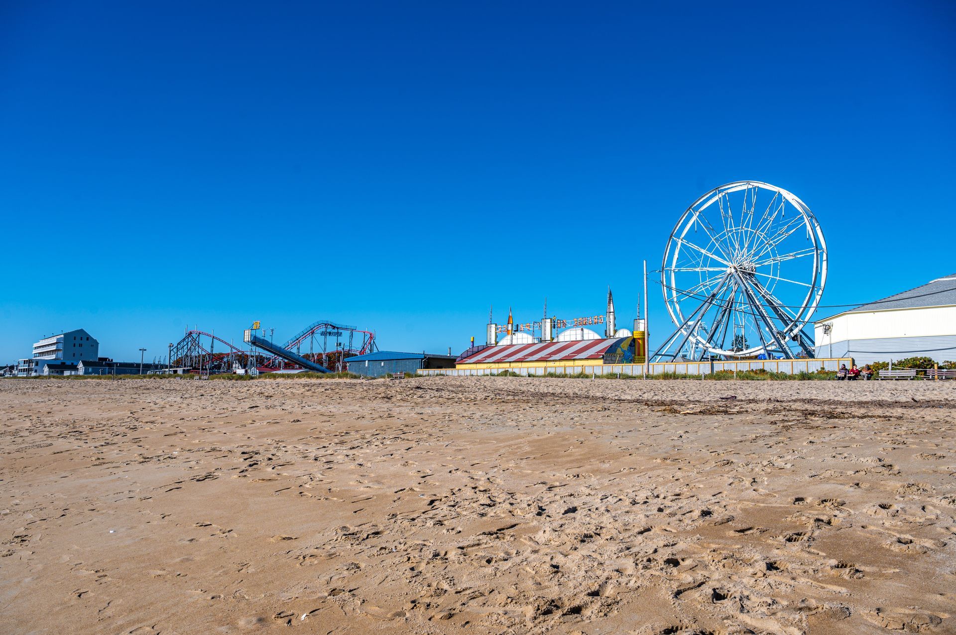 A sandy beach leads toward a boardwalk amusement park featuring a large Ferris wheel and roller coaster under a blue sky.