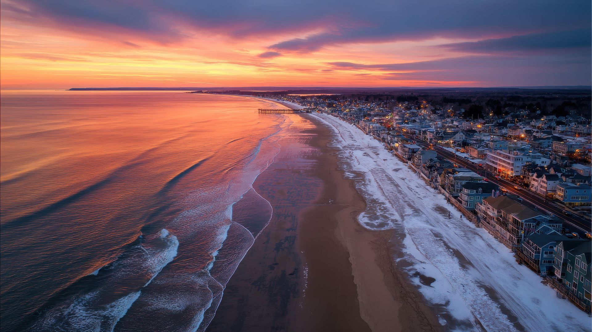 An aerial view of a sandy beach at sunset, with snow lining the shoreline and a town situated along the coast.