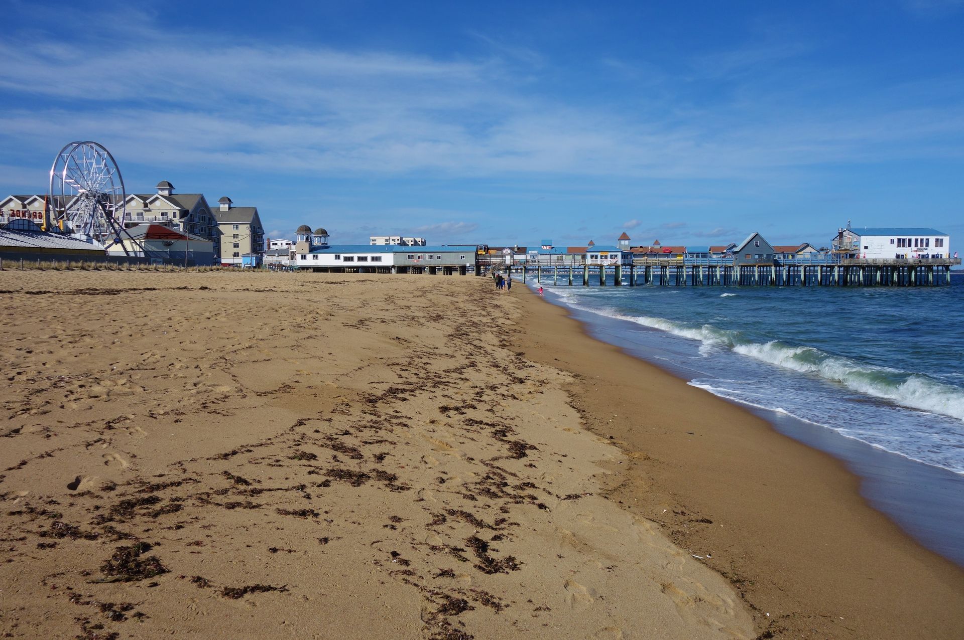 A sandy beach leading to a pier and a Ferris wheel under a clear blue sky.