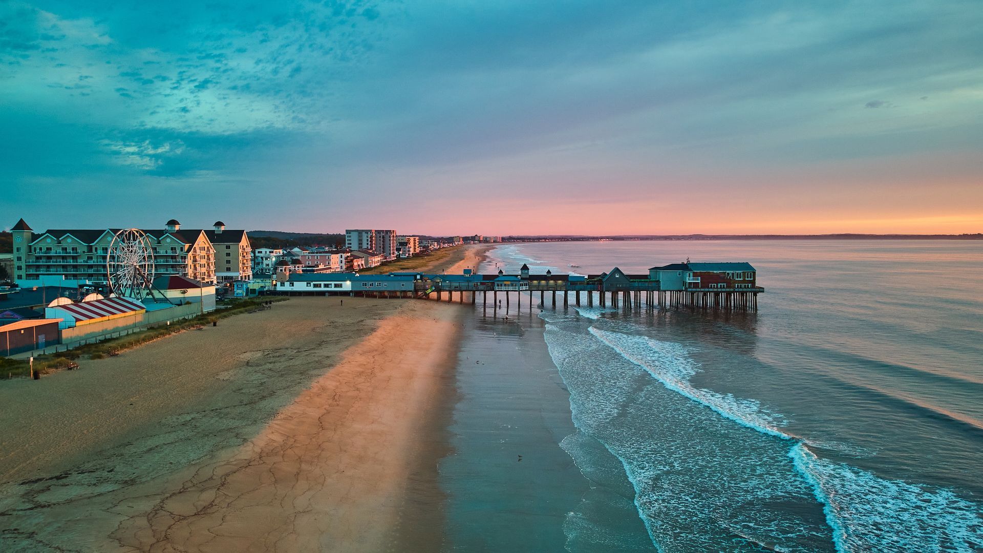 An aerial view of an ocean pier and sandy beach at sunrise with a glowing, colorful sky and nearby buildings.