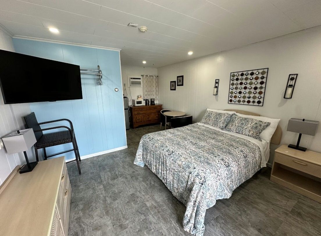 A tidy hotel room features a queen bed, light blue accent wall with a wall-mounted TV, and light wood furniture.