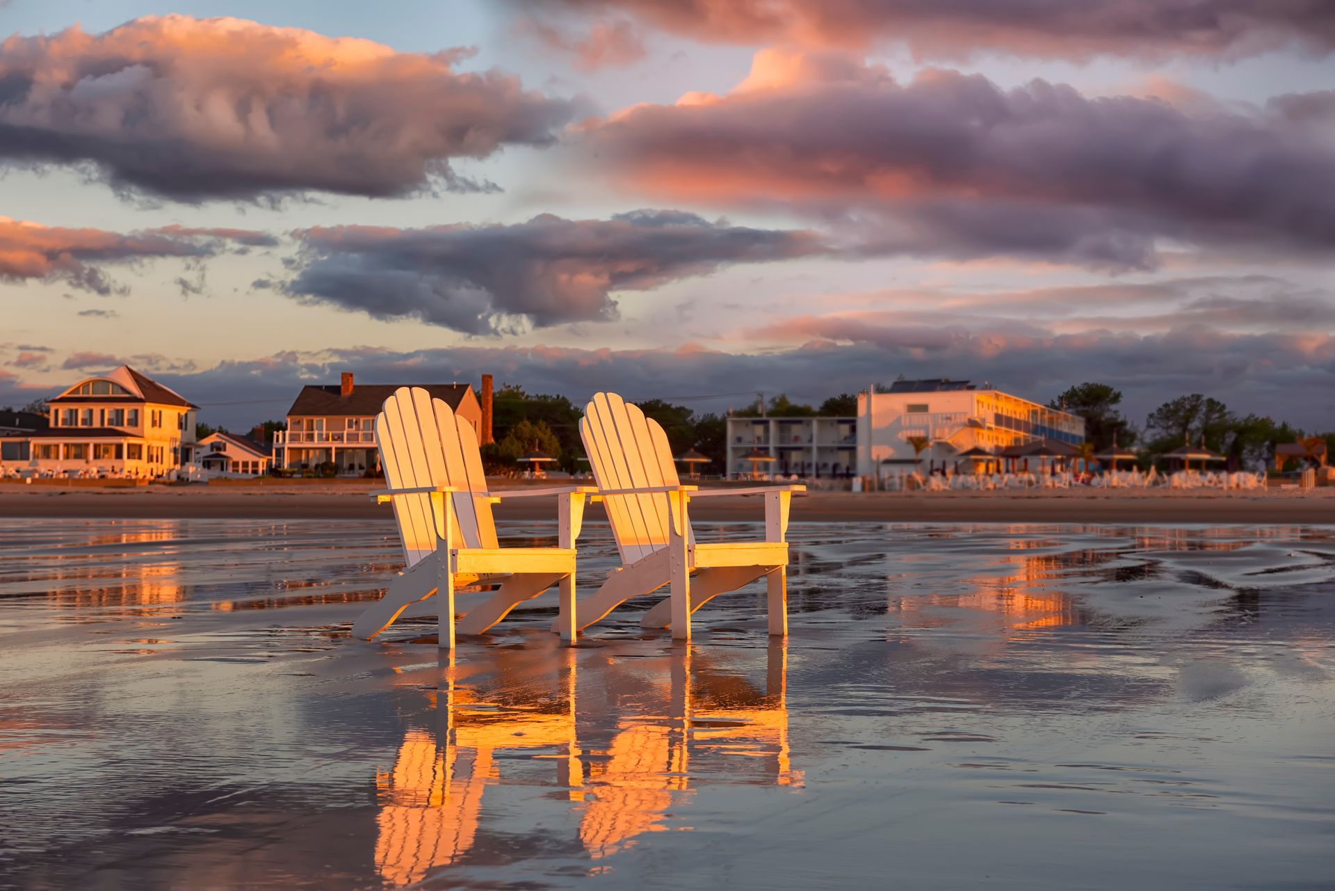 Two white wooden beach chairs sit on a wet sandy shore reflecting the warm, golden colors of a sunset sky.