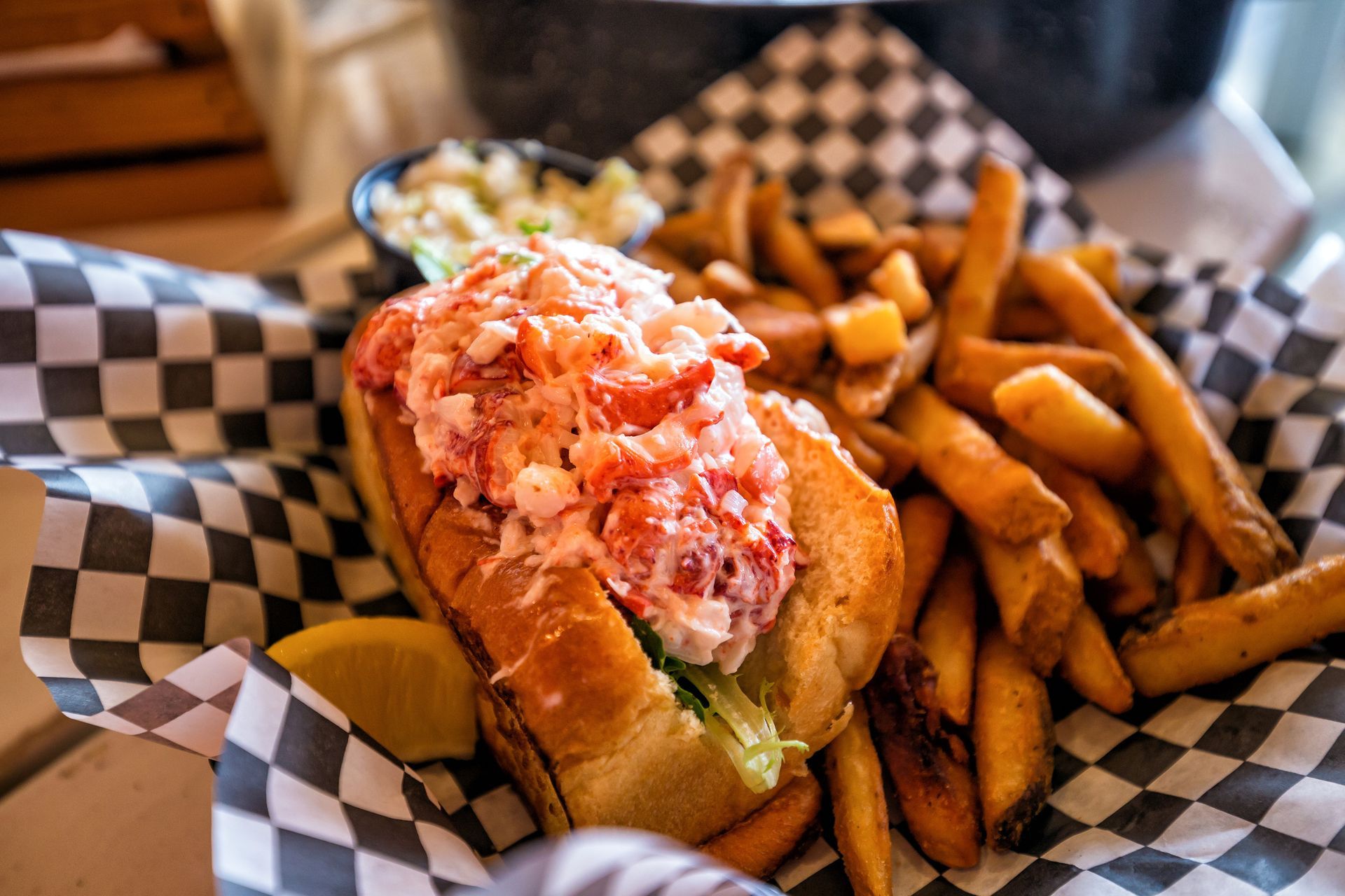 A lobster roll with a side of fries and coleslaw served in a basket lined with black and white checkered paper.