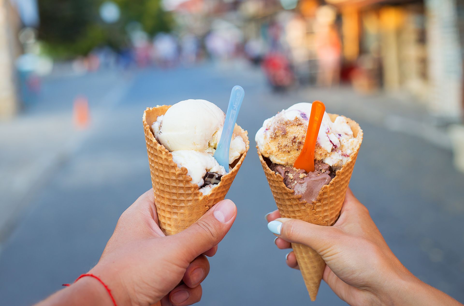 Two hands hold ice cream cones with colorful spoons against a blurred street background.