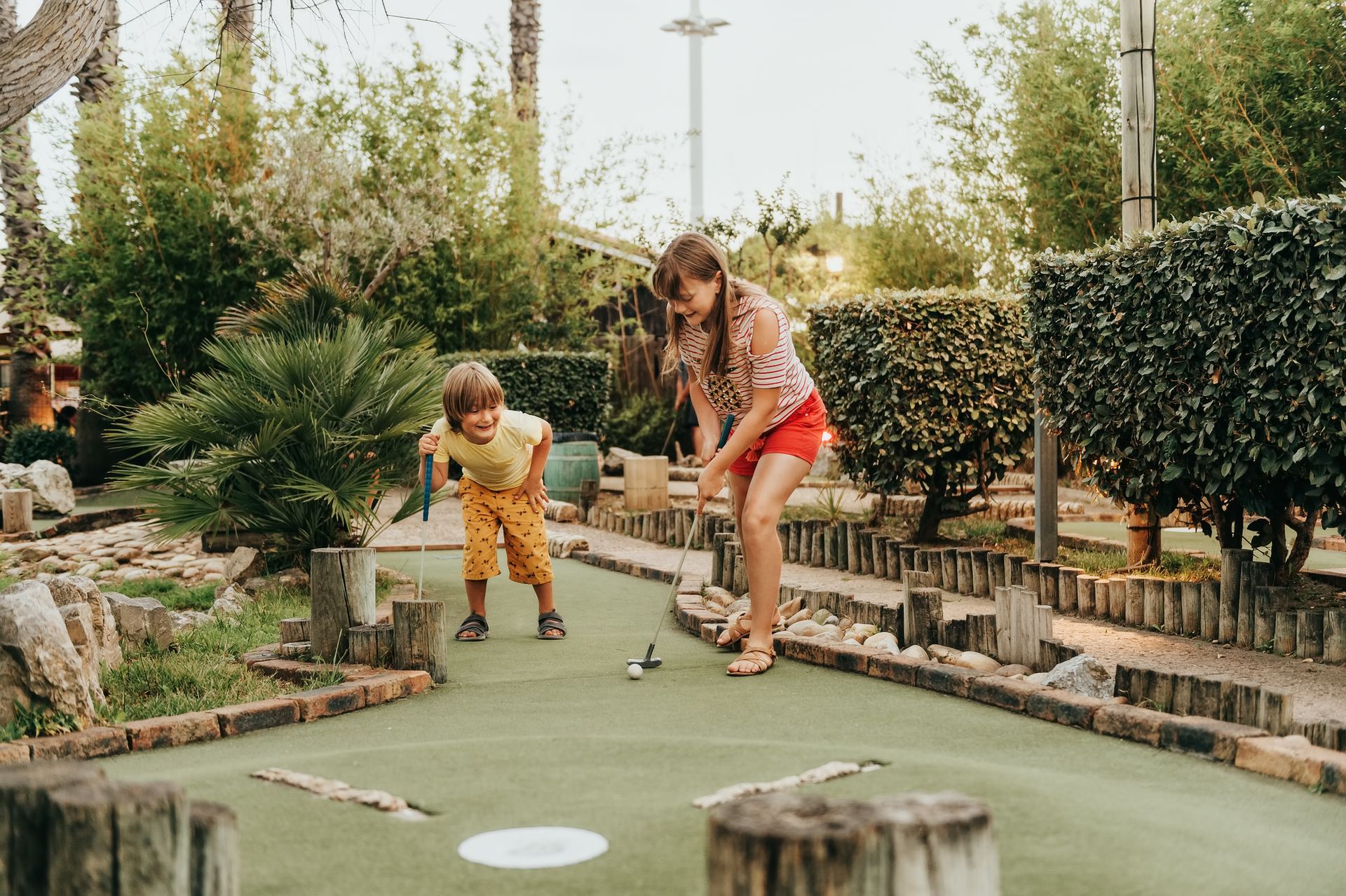 Two children play miniature golf on an outdoor green course surrounded by bushes and trees.