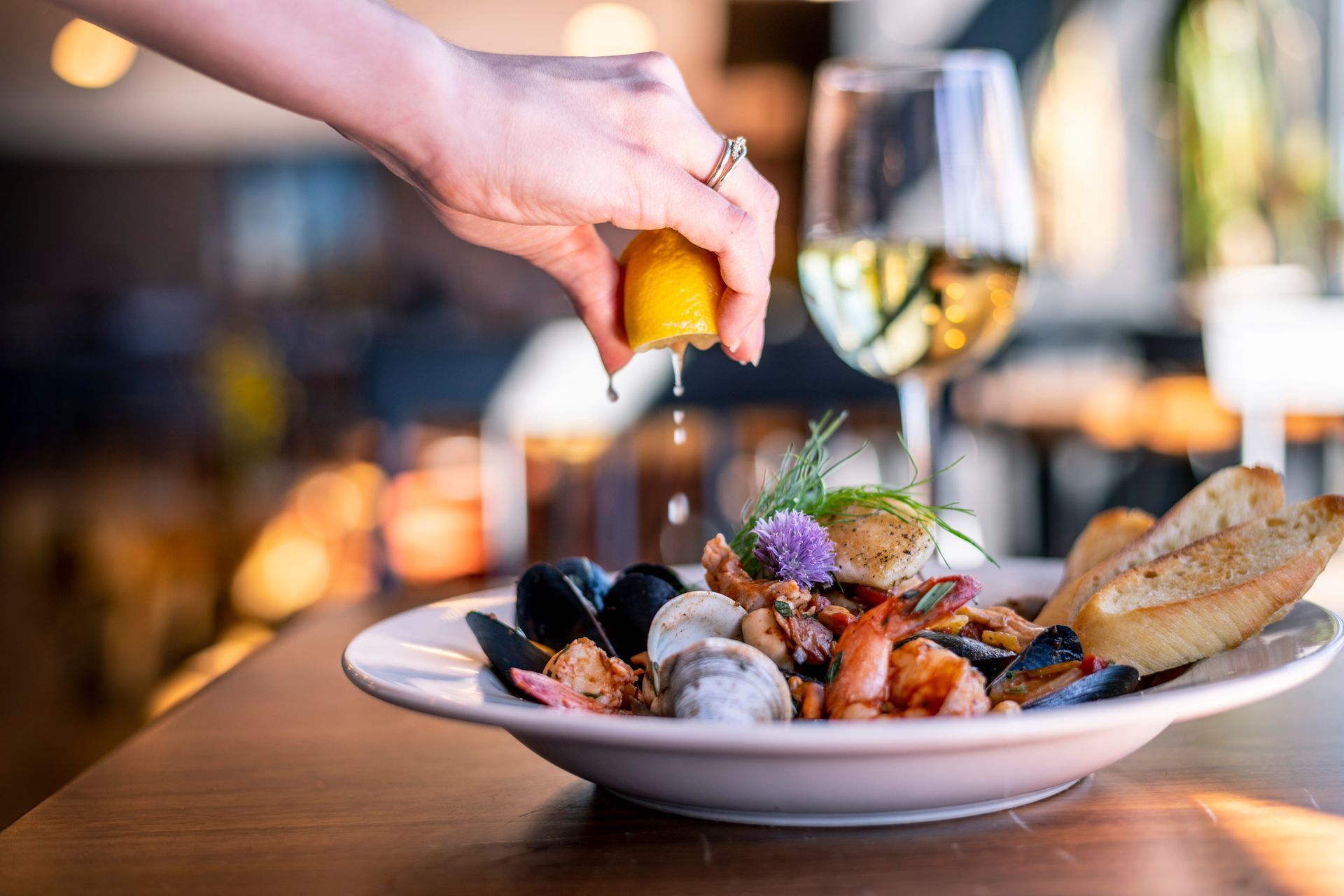 A hand squeezes a lemon wedge over a bowl of mixed seafood and toasted bread, with a glass of white wine in the background.