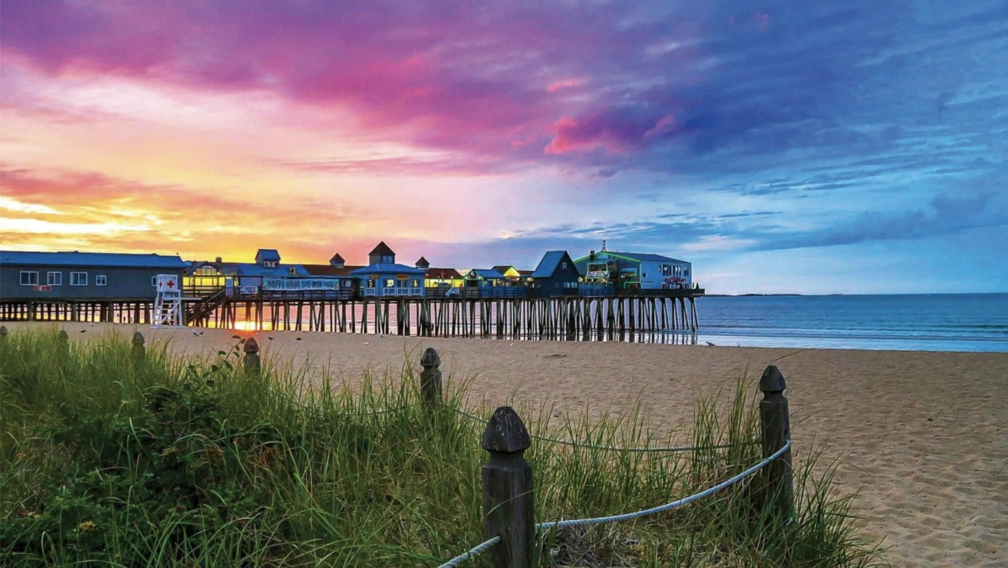 A beach pier extends into the ocean at dusk, with a vibrant sky transitioning from purple and orange to deep blue.