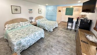 A hotel room with two beds, light blue accents, a small kitchen area with a high chair, and a television on a dresser.