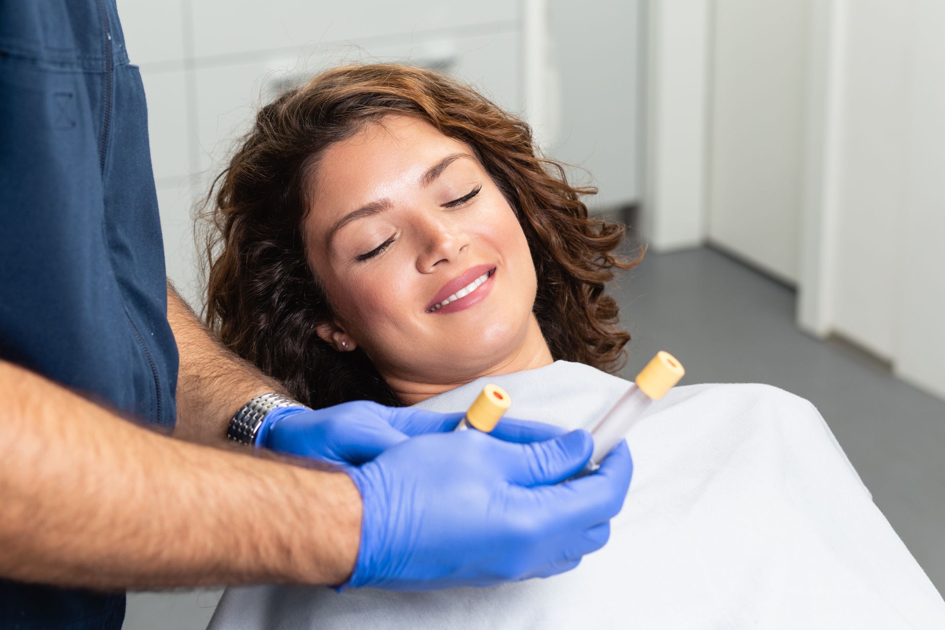 A woman is smiling while sitting in a dental chair while a dentist is holding a syringe.