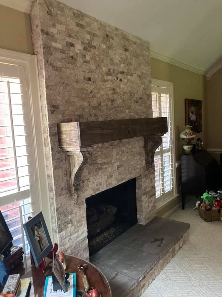 Fireplace with stone facade and dark wood mantel, flanked by windows with white shutters.