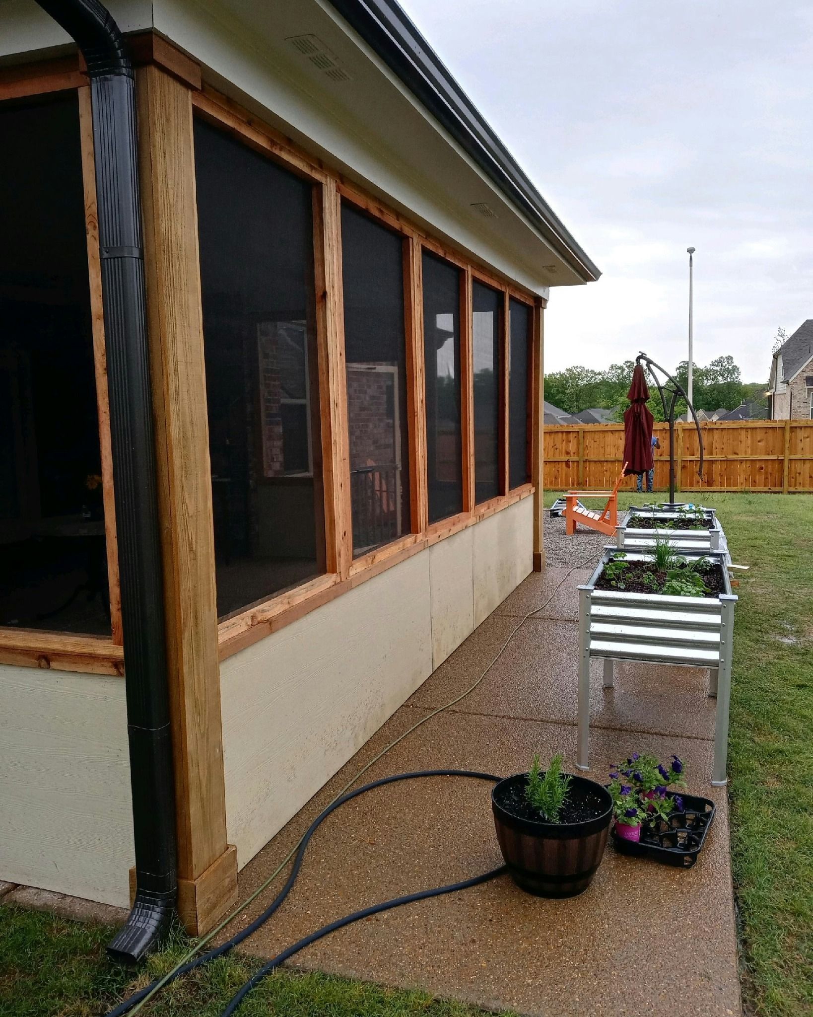 Screened porch with wooden frames, next to a garden and a lawn, with rain water on concrete.