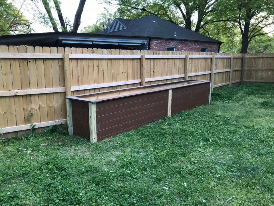 A long, brown raised garden bed against a wooden fence in a grassy backyard.