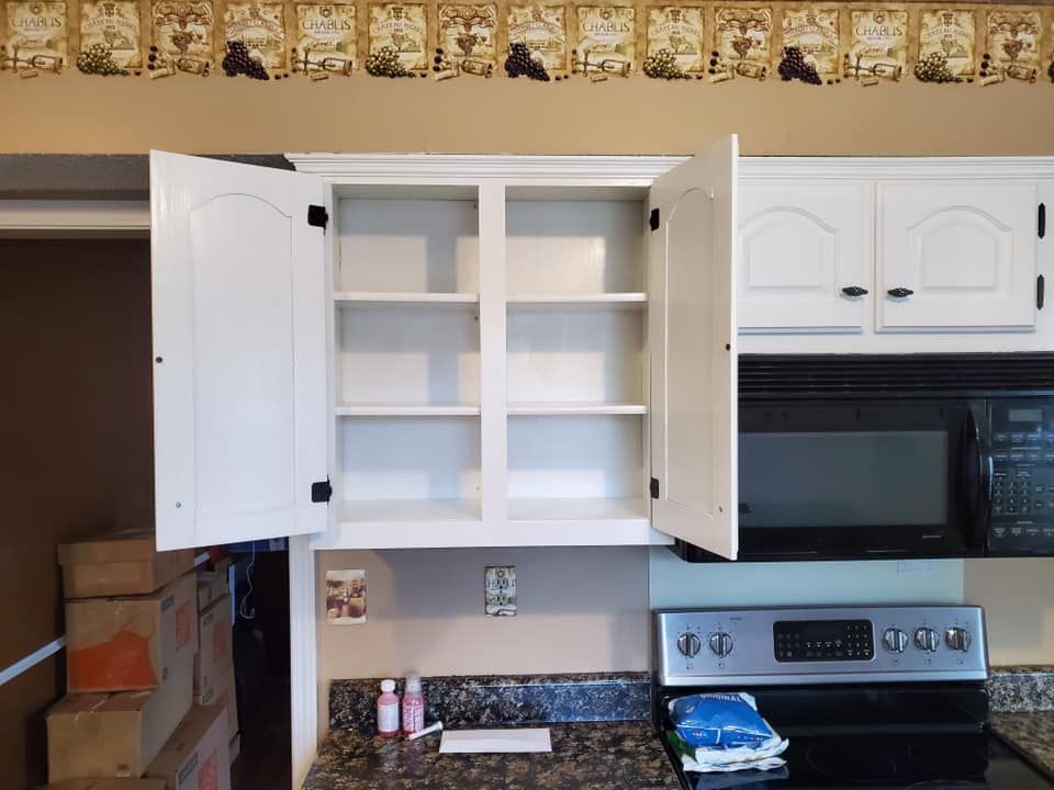 White kitchen cabinets, one open to reveal shelves, above stove and microwave. Brown countertop and beige wall.