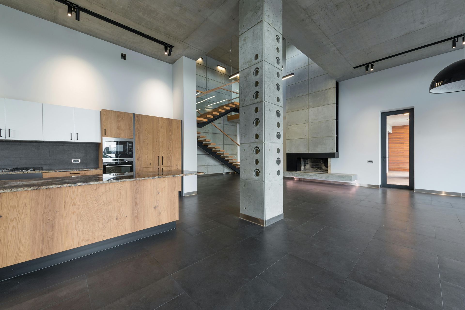 Modern open-plan living space with kitchen, fireplace, and staircase. Black flooring, concrete pillars, and wood accents.