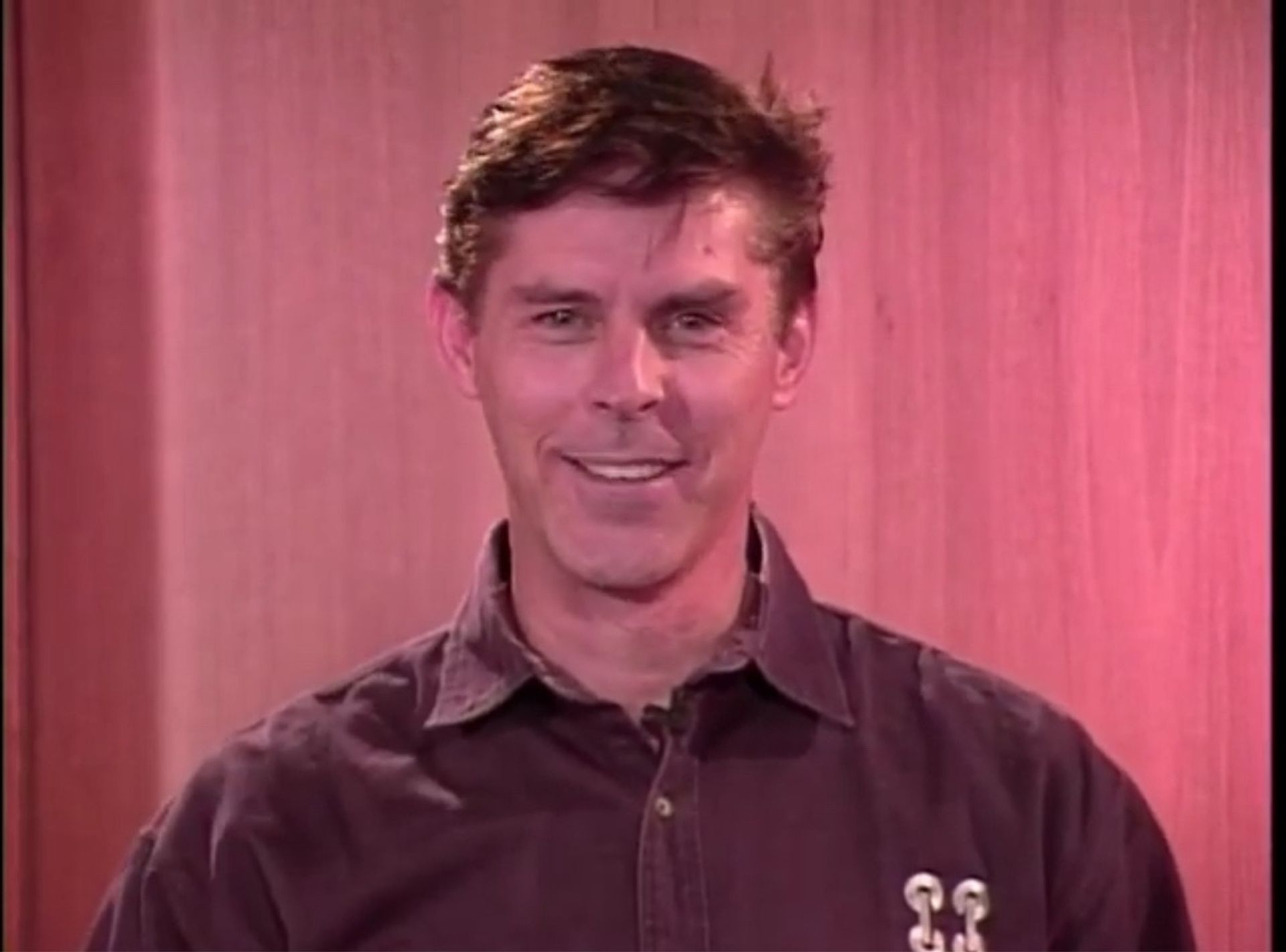 Tom Wright in a purple shirt is smiling in front of a pink wall.