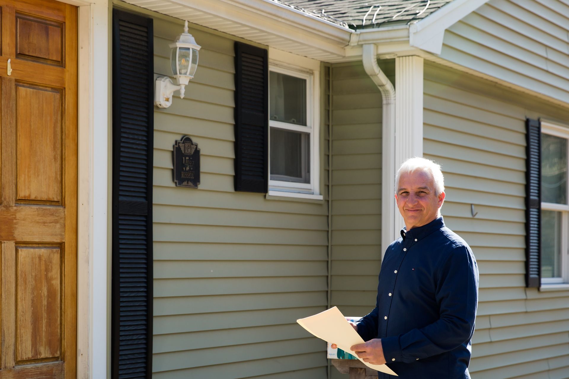 A man is standing in front of a house holding a piece of paper.