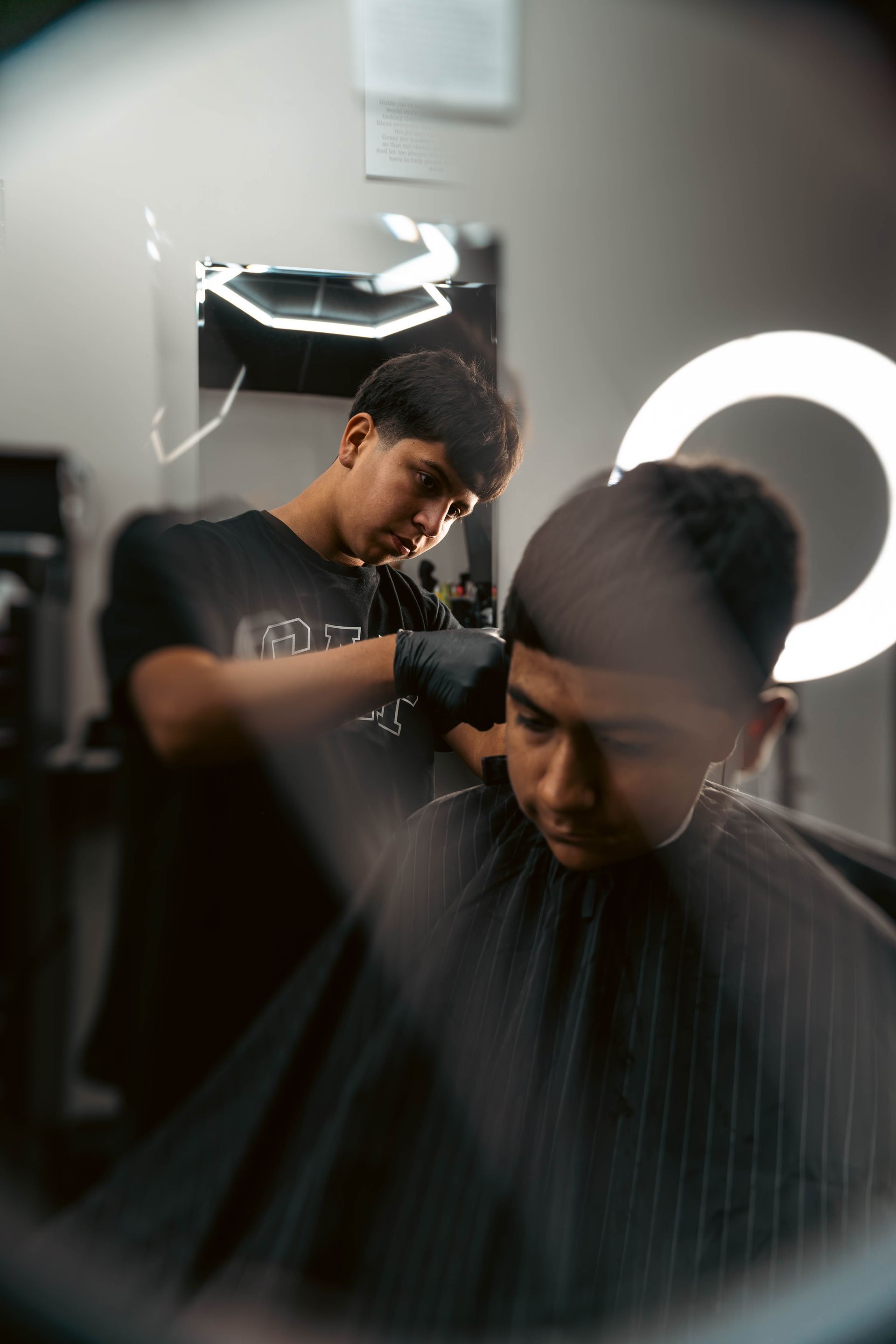 A barber wearing black gloves performs a haircut on a client in a studio, captured through a circular lens reflection.