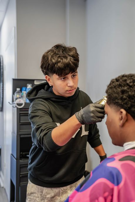 A barber wearing a black hoodie and gloves uses electric clippers to groom a client wearing a pink and blue patterned cape.