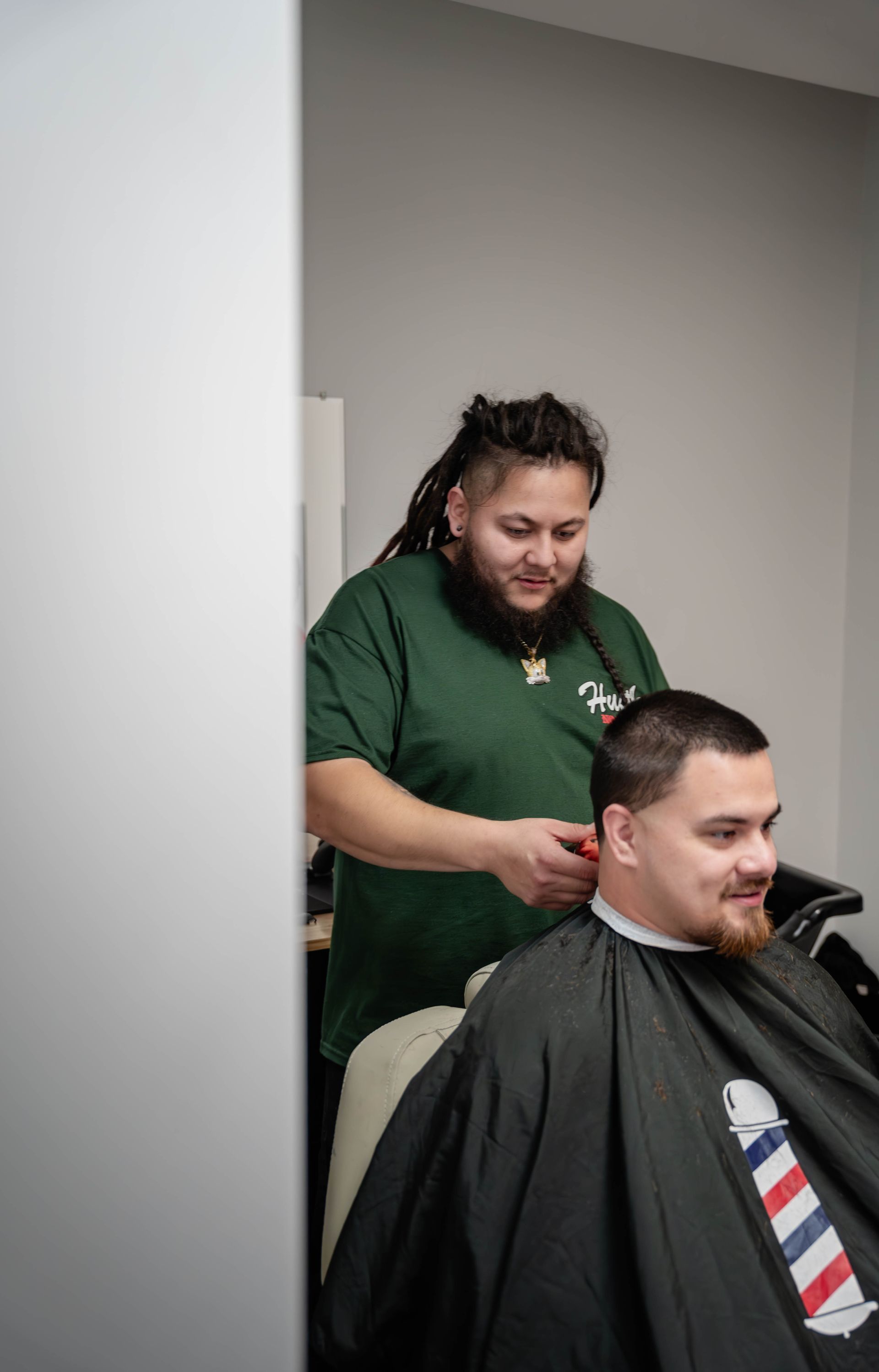 A barber with dreadlocks wearing a green t-shirt trims the hair of a customer in a black salon cape.