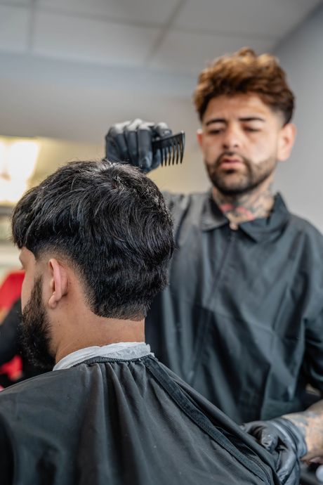 A barber wearing black gloves uses a comb to trim the hair of a client wearing a black cape in a salon.